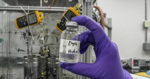 Researcher in purple gloves holding a hand labeled sample bottle in front of laboratory equipment