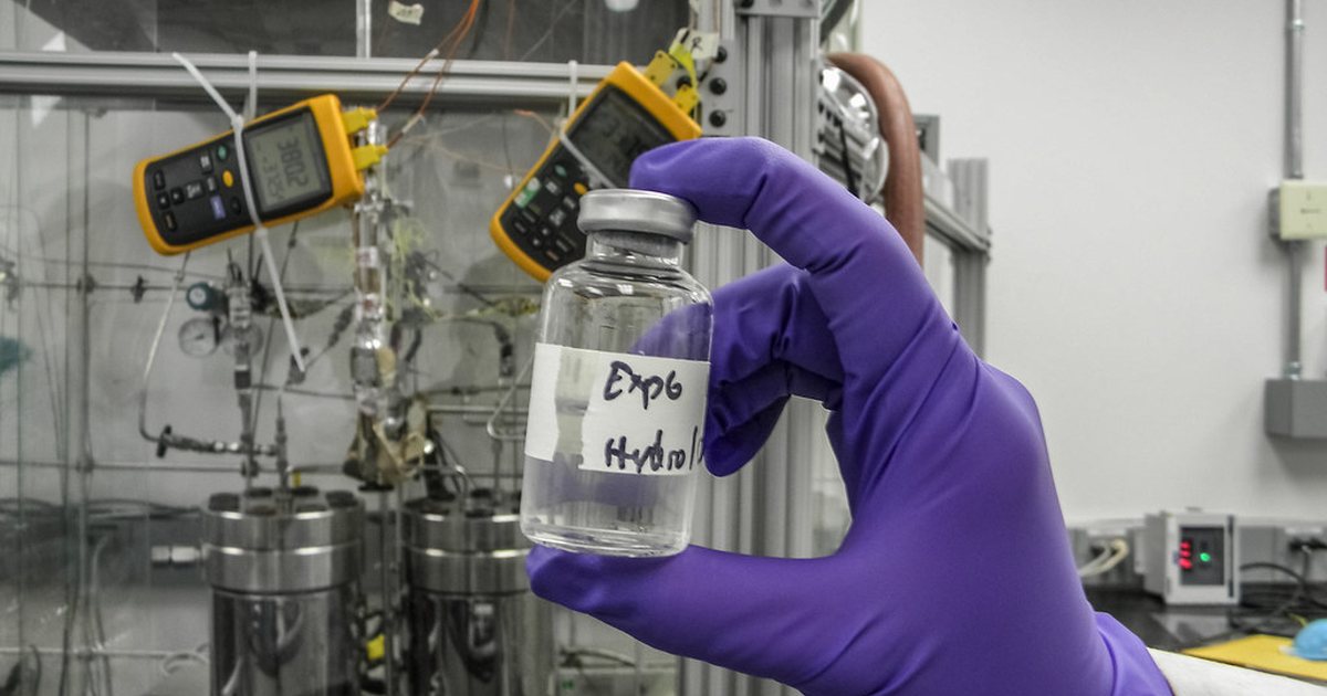 Researcher in purple gloves holding a hand labeled sample bottle in front of laboratory equipment