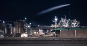 Large industrial processing plant illuminated against a black night sky with a plume rising from a stack