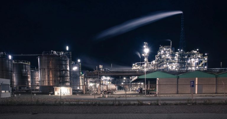 Large industrial processing plant illuminated against a black night sky with a plume rising from a stack