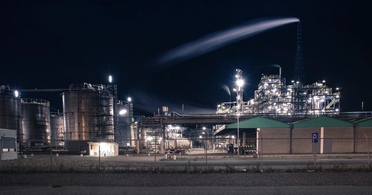 Large industrial processing plant illuminated against a black night sky with a plume rising from a stack