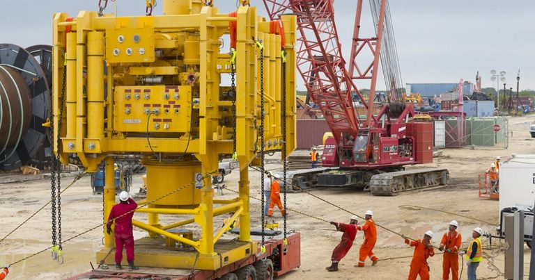 Crew in orange coveralls and hard hats guiding a large piece of yellow subsea oilfield equipment with a crane