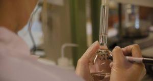 Gloved hands labeling a glass laboratory flask containing liquid on a bench